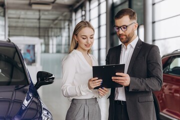 Car Sales Manager Showing Auto To Caucasian Lady Buyer Standing In Luxury Automobile Dealership Store. Buying Vehicle Concept