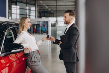 Young caucasian woman buys a car at a car dealership. Happy smiling bearded man seller gives the car keys