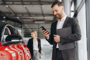 Smiling salesman using smartphone while having business call at car showroom. Car dealer holding...