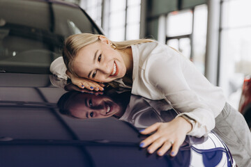 Positive young caucasian woman hugging his new car in dealership. Happy woman finally gets...