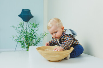 A 9-month-old baby boy in a stylish shirt, pants and sneakers is standing near a support.