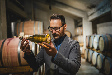 Adult man winemaker hold and check bottle of wine between barrels