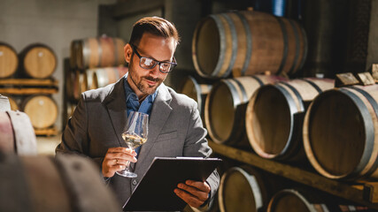 Adult man winemaker hold glass stand between the barrels in cellar