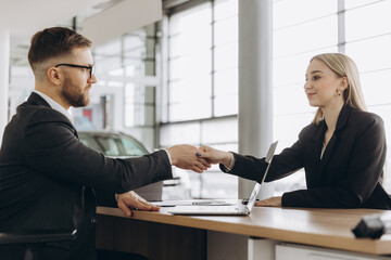 A bearded man in a suit, a manager, a car salesman in a car dealership, who signs a contract at the table with a woman buyer and shakes her hand