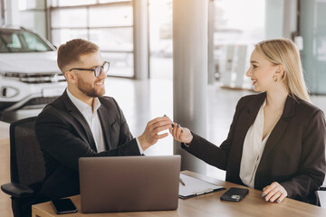 Sitting by the table and taking the keys. Man with woman in white clothes are in the car dealership together. Car sales contract