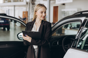 Handsome car dealership female worker in suit is holding a folder and smiling while standing near...