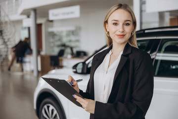 Handsome car dealership female worker in suit is holding a folder and smiling while standing near...