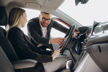 Friendly male manager showing young female customer cars. Beautiful smiling woman sitting inside vehicle in car. Bearded sales consultant working in car dealership.