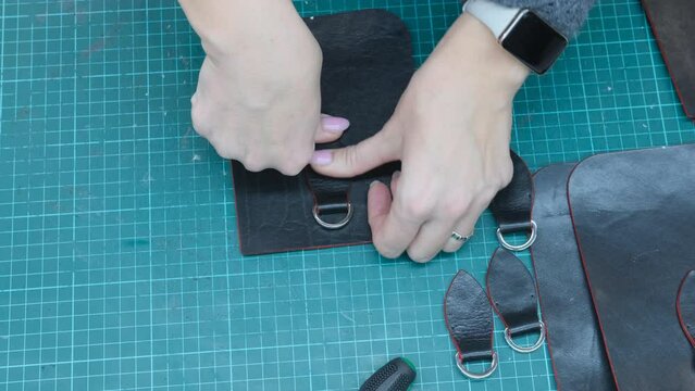 The craftsman carefully assembles a black leather bag, gluing the strap on a green cutting mat in a well-lit workshop.