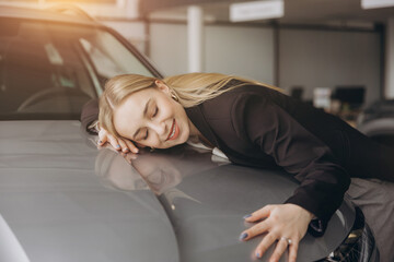 Positive young caucasian woman hugging his new car in dealership. Happy woman finally gets...