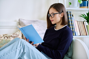 Young woman reading book, sitting on couch at home, engrossed in fiction