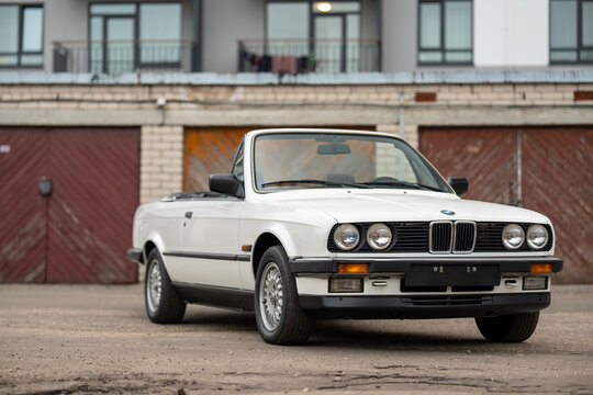 Riga, Latvia - April 1, 2024: The white classic convertible BMW E30 car with opened roof. An old garages and modern building on the background.