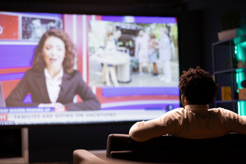 Man sitting on couch in front of ultrawide smart TV displaying news program broadcasting. Person watching big television screen showing VOD channel with newscast coverage