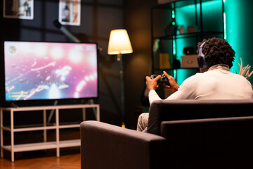 Man plays science fiction singleplayer game on smart TV while talking with friends through headphones. Player enjoying videogame with high quality graphics, using controller to navigate spaceship