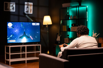 BIPOC man in home theatre using cloud gaming service to play demanding science fiction videogame on smart TV. Gamer enjoying high quality game, streaming it online, flying spaceship with controller