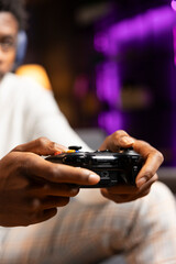 Close up shot of gamepad controller in BIPOC man hands playing intense online videogame in neon illuminated living room. Gamer participating in internet esports competition, playing on gaming console