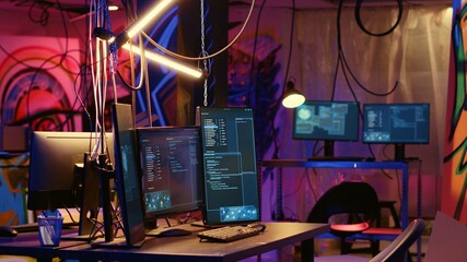 Programming language on PC screens in empty messy hackers base of operations with grunge graffiti drawings sprayed on walls. Neon lit ghetto hideout used by criminals to commit illegal activities