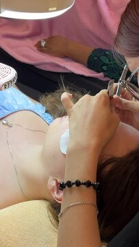 Video of process of eyelash extension, young lashmaker sitting over client lying on couch and gluing fake lashes on lid using tweezers. Concentrated master holding tools and touching cilia.