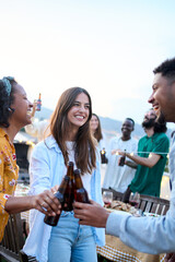Vertical. Young group of multi ethnic young people dancing together and toasting beer at rooftop party outside. Happy friends having fun cheering drink while celebrating birthday at home terrace.