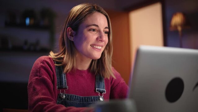 Young smiling Caucasian woman in front of laptop in living room. Beautiful blonde using computer to chat online with friends. Millennial female typing on social apps on electronic device at night home