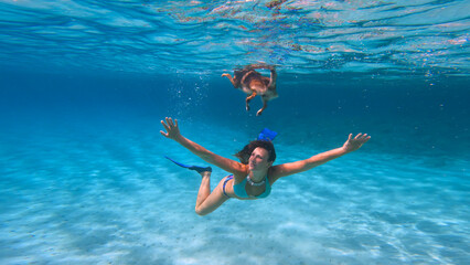 UNDERWATER: Beautiful woman in bikini dives under her dog that swims on the surface of crystal clear sea. Joyful moment between a young lady and her doggo swimming together in blue Adriatic Sea.