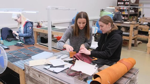 Two women collaborate on designing and crafting leather handbags in a well-organized and busy workshop, focusing on precision and detail in their handmade creations.