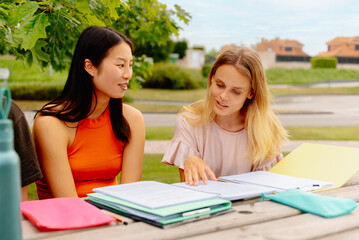 Two girls, one Asian and one white, studying together with class notes sitting on a bench outside the university campus. Students preparing for an exam together.