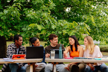 multiracial and diverse group of students working together with laptops and class notes on a common project sitting at a table outside the university campus.
