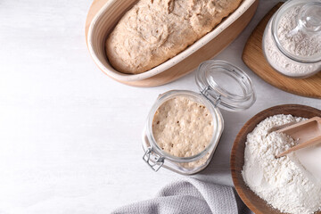 Sourdough starter in glass jar, flour and dough on light table, flat lay. Space for text
