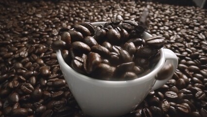 Close up of coffee bean falling in to coffee cup surrounded by pile of beans. Dropping aromatic...