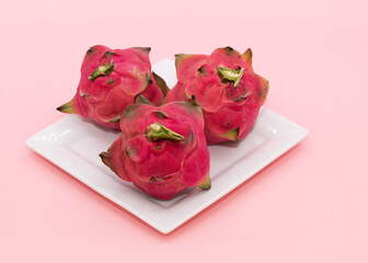 Three dragon fruits with pink skin and green spikes are on a white plate against a light pink background.