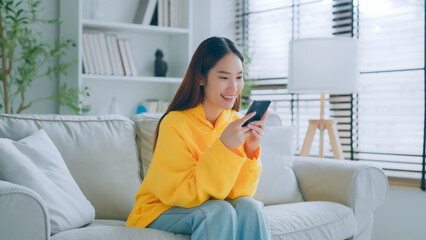 Happy young asian woman relax on comfortable couch at home texting messaging on smartphone, smiling girl use cellphone chatting, online shopping at home, video call communication