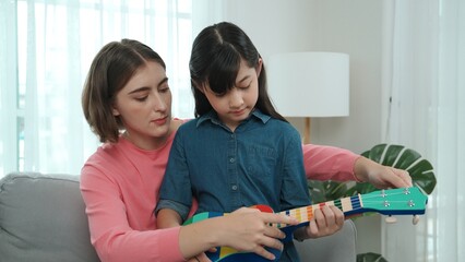 Happy girl playing ukulele while caucasian mom teaching and explain about acoustic music at home....