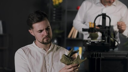 A young guy research assistant holds in hands and views khaki coloured plastic model of human mandible. A middle aged man mechanical engineer uses digital calipers and customize the work of 3d printer