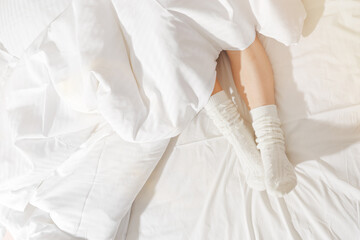 Morning cozy mood, Time to relax, Top view woman legs in knitted white socks under blanket on bedclothes, legs close up, aesthetic lifestyle, empty space for text, sunlight glare, Home weekend
