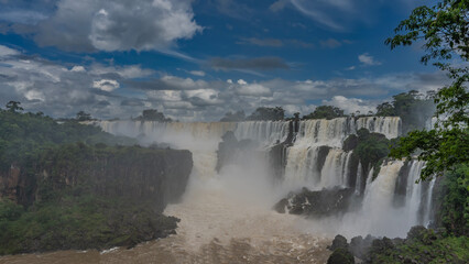 A beautiful cascade of waterfalls. White foaming streams collapse from the rocks into the riverbed. Spray, fog rising. Clouds in the blue sky. Lush green vegetation. Iguazu Falls. Argentina.