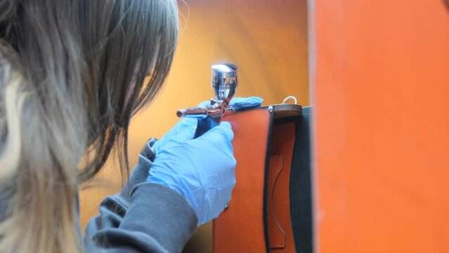 A leatherworker uses an airbrush to add finishing touches to a brown leather bag, highlighting the precision and detail of this craft.