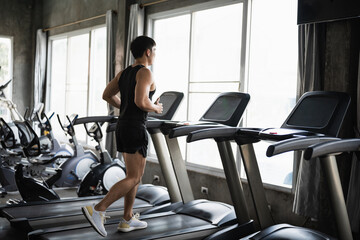 A man in a black tank top and shorts runs on a treadmill in a modern gym, surrounded by various...