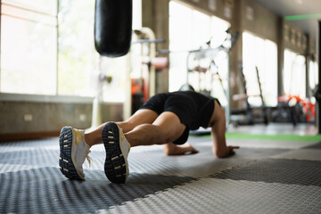 A focused man in a black tank top and shorts holds a plank position on a gym floor. The well-lit,...