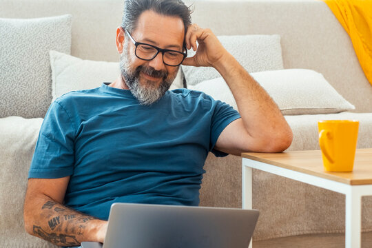 Adult man in casual clothing and eyeglasses using laptop and smiling while sitting on the floor against the sofa at home. Indoor technology computer leisure activity people lifestyle. Working online
