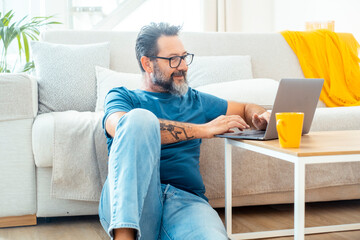 Adult man in casual clothing and eyeglasses using laptop and smiling while sitting on the floor...