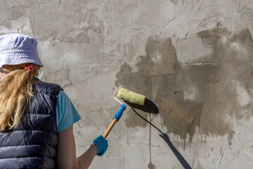 A woman in a blue vest, blue shirt and hat primes a textured concrete wall with a roller and brush....