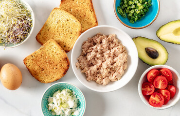 Set ingredients for cooking healthy sandwich with avocado, tuna, eggs, cherry tomatoes and fresh microgreens on white marble background top view