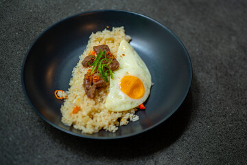 beef saikoro rice bowl and sunny side up egg on black plate, over stone tile