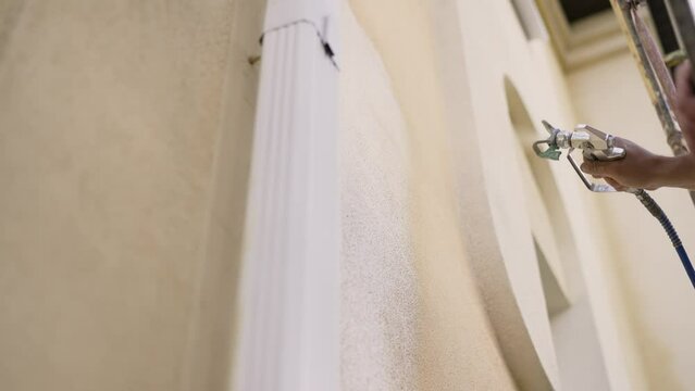 Hand of a contractor using a spray gun to paint the plaster of a house, close-up. A man's hand holds a spray gun, uses a chemical solution, paints the house. Painting the outside of a house with a spr