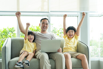 Asian father and his two children cheerfully using a laptop together on the couch in their bright,...