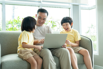 Asian father and his two sons have fun watching video in laptop while sitting on sofa in living room at home. Family relaxing on holiday.