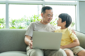 Asian father and son have fun watching video in laptop while sitting on sofa in living room at home. Family relaxing on holiday.