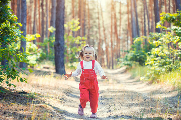 Portrait of smiling three years old caucasian child girl runs on the trail in the pine forest. Waving hands. Forest on bright light background.