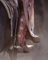 western cowboy boot with silver spur on cowgirl colored stylish boot with pattern and design on leather sitting on horse chinks rope and saddle pad visible close up vertical image room for type 